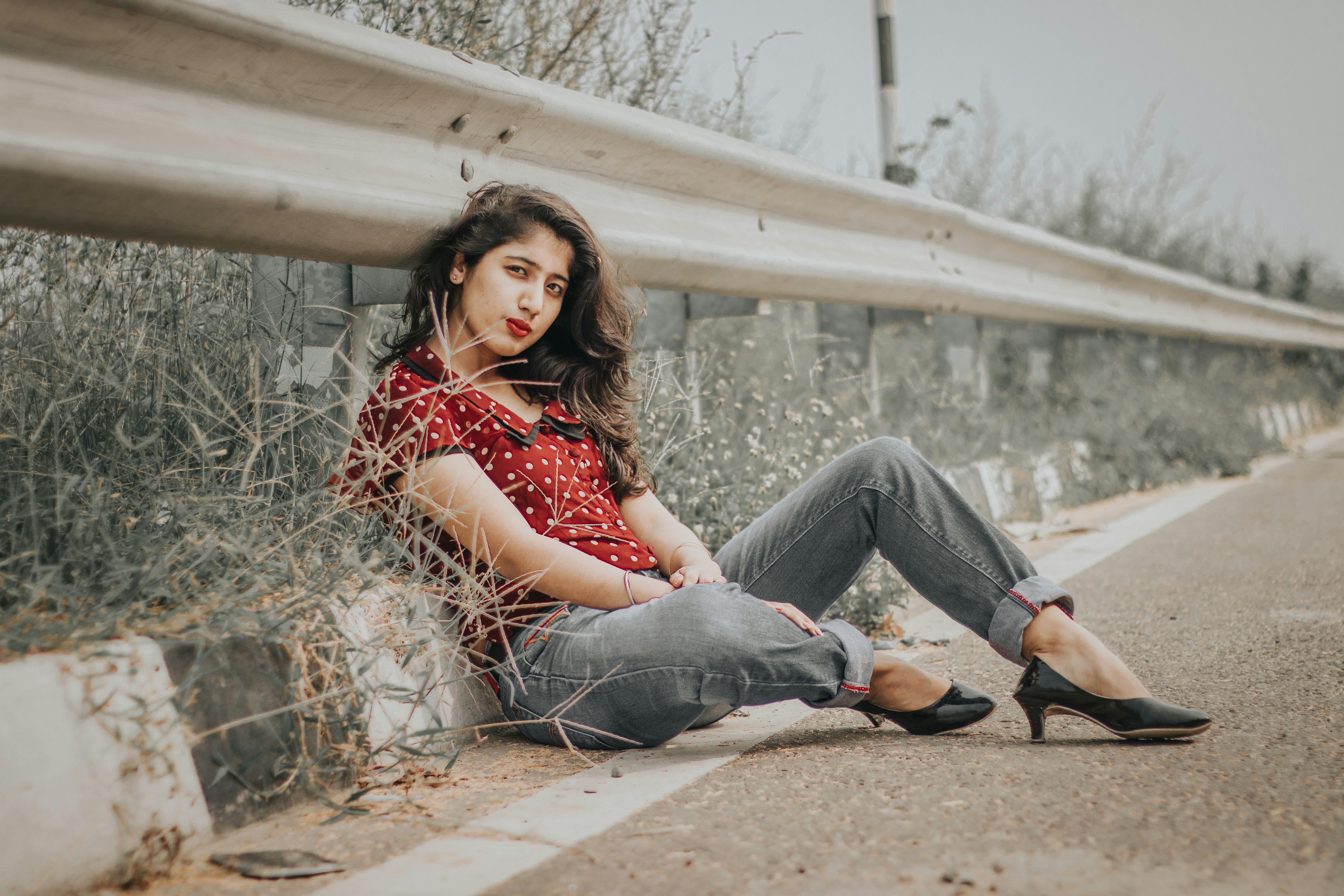 woman in red dotted shirt and gray denim pants sitting on ground near road rail